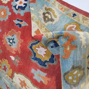 Op de afbeelding: Opgerolde tapijt met een rode achtergrond en ingewikkelde patronen in blauw, oranje en geel. De rand is lichtblauw. Het ontwerp suggereert een traditionele of vintage stijl, met aandacht voor details.