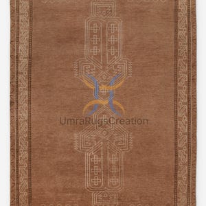 Peut inclure: Tapis rectangulaire marron avec un motif géométrique central blanc cassé et bleu. Le tapis présente une bordure avec des motifs répétitifs. Le texte "UmraRugsCreation" est visible.