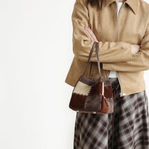 Peut inclure: Un sac fourre-tout patchwork avec une poignée en cuir marron. Le sac présente un mélange de patchs en daim marron, beige et crème. La personne porte une veste en daim beige et une jupe à carreaux.
