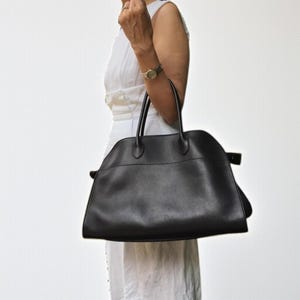 Puede incluir: Un bolso de mano de cuero negro con dos asas superiores. El bolso tiene una forma estructurada con una parte superior curvada y un sutil detalle de diseño en el lateral. El bolso lo lleva una persona que lleva un vestido blanco.