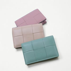 Może przedstawiać: Trzy skórzane portfele w pastelowych kolorach: różowym, beżowym i turkusowym. Każdy portfel ma tkany wzór z przodu. Prostokątne portfele wydają się zamknięte, co sugeruje, że są przeznaczone do noszenia kart lub gotówki.