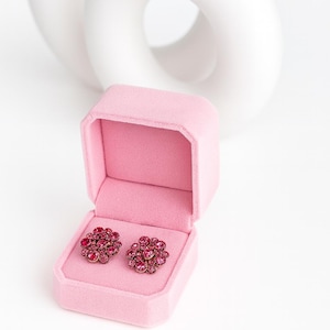 May include: A pair of ruby-colored floral earrings displayed in an open pink velvet jewelry box. The earrings are made of small, sparkling stones arranged in a flower shape. A white, abstract sculpture is in the background.