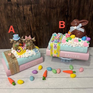 May include: Two Easter-themed decorations. One features two Highland cows with glasses and hats, atop pastel books with the words "HOPPY EASTER." The other has a chocolate bunny and colorful eggs. Decorative carrots and eggs are scattered around.