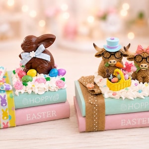 May include: Two decorative stacks of books labeled HOPPY and EASTER. The left stack features a brown chocolate-style bunny with a blue bow. The right stack features two highland cows with glasses, a top hat, a pink bow, and a yellow basket.