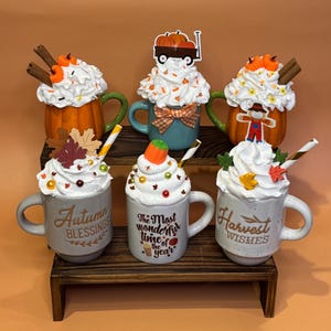 May include: A collection of decorative mugs with whipped cream toppings and fall-themed decorations. The mugs are arranged on a wooden shelf, featuring pumpkins, leaves, and the phrases "Autumn Blessings", "The Most Wonderful Time of the Year", and "Harvest Wishes".