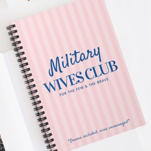 May include: A spiral-bound notebook with a pink and white striped cover. The text "Military Wives Club" is in blue script, with the phrase "For the Few & The Brave" below. The bottom of the cover reads "Drama included, wine encouraged."