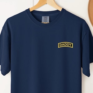 May include: Navy blue t-shirt with the word "DADDY" in a yellow rectangular box on the chest. The shirt is hanging on a wooden hanger. The brand label "Comfort Colors" is visible on the back of the neck.