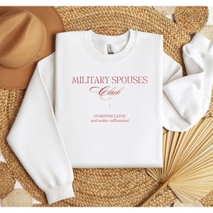 May include: White crewneck sweatshirt with the text "MILITARY SPOUSES Club" in burgundy script. Below the club name is the phrase "OVERSTIMULATED and under caffeinated" in a simple font. The sweatshirt is displayed on a woven surface.