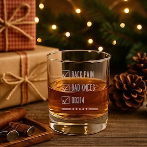 以下が含まれることがあります： 「BACK PAIN」、「BAD KNEES」、および「DD214」の文字が刻まれた透明なウイスキーグラス。グラスには琥珀色の液体が入っています。背景には、包装されたギフト、松ぼっくり、ストリングライトがあります。