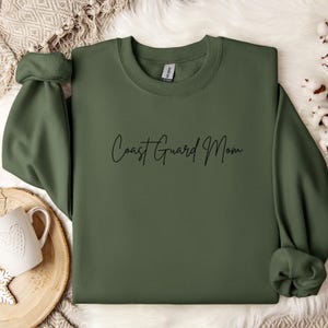 May include: An olive green sweatshirt with the words "Coast Guard Mom" in black script. The sweatshirt is made of a soft material and has a crew neck. The sleeves are cuffed. The sweatshirt is displayed on a white surface.