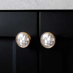 May include: Two decorative cabinet knobs with a gold-colored rim and a faceted, clear, and white center. The knobs are mounted on a black cabinet door. The knobs add a touch of elegance to any cabinet.