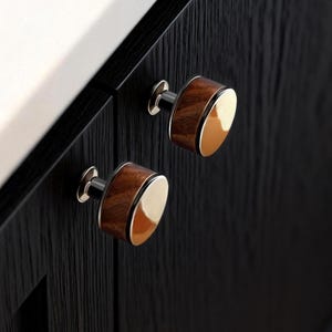 Puede incluir: Primer plano de dos tiradores de armario. Cada tirador presenta un centro de veta de madera oscura con una cara redondeada de color dorado. Los tiradores están montados en una puerta de armario negra con una textura de veta de madera visible. La ferretería tiene una base plateada.