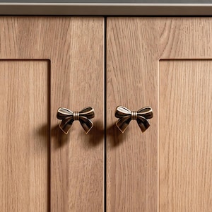 Op de afbeelding: Close-up van een houten kast met twee bronskleurige strikvormige handgrepen. De kastdeuren zijn gemaakt van lichtgekleurd hout met zichtbare nerf. De handgrepen zijn symmetrisch en geven de kast een decoratieve uitstraling.