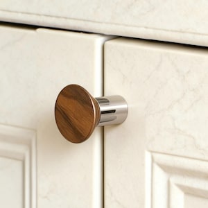 May include: A cabinet knob with a round, wooden face and a polished chrome base. The wood grain is visible on the face of the knob. The knob is attached to a white cabinet door with a marble-like pattern.