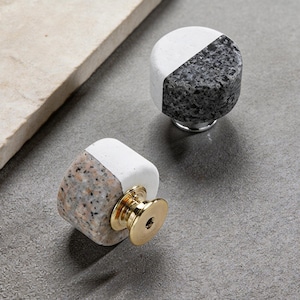 May include: Two decorative cabinet knobs. Each knob is a rounded rectangle, split diagonally with a white and speckled stone finish. The knobs have a gold-colored base and are set against a gray background.