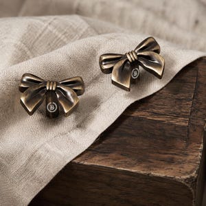 May include: Two bow-shaped cabinet knobs crafted from antique bronze-toned metal. Each knob features a central knot detail and a small circular emblem on the base containing the letter B surrounded by a decorative laurel wreath border.