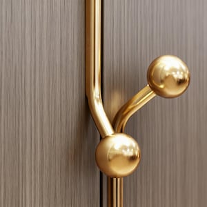 May include: A gold-toned metal coat hook featuring a vertical bar with two protruding arms, each ending in a smooth, spherical knob. The hardware is mounted against a vertical, light brown wood-grain surface with a visible linear texture.