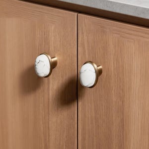 Puede incluir: Dos tiradores de armario con caras ovaladas blancas con aspecto de mármol y bases de metal color latón. Los tiradores están montados en una puerta de armario de madera marrón claro. Herrajes decorativos.