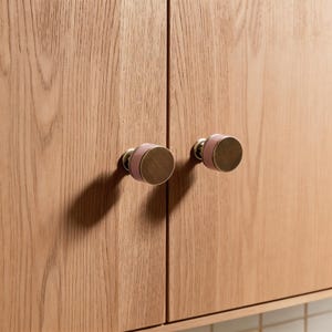 Puede incluir: Primer plano de dos pomos de armario. Cada pomo tiene una base circular de color latón con un acento rosa y un tirador circular a juego. Los pomos están montados en una puerta de armario de madera de color claro.