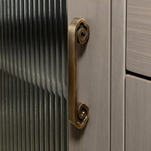 Può includere: Primo piano di un'anta di armadio con una maniglia color ottone. La maniglia ha un design decorativo a forma di nuvola a ciascuna estremità. L'anta dell'armadio è di colore grigio chiaro.