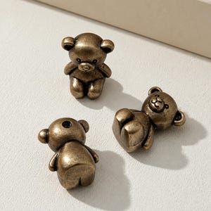 May include: Three small, three-dimensional teddy bear figurines crafted from a dark, antique-toned metal. Each bear features a smooth, rounded head, small ears, and a seated posture. One bear is positioned upright, while two others lie flat.