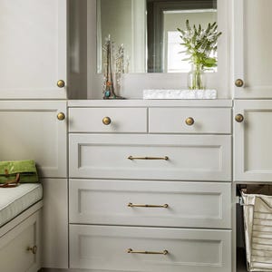 May include: A light grey fitted cabinet with drawers and a bench seat. The drawers have brass handles and knobs. A mirror and decorative items sit on top of the cabinet. A green bag rests on the bench.