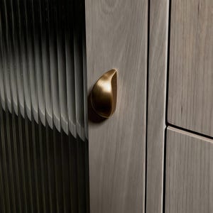 May include: A light gray wood cabinet door featuring a vertical fluted glass panel and a teardrop-shaped brushed brass handle. The handle is mounted on the wooden frame, which displays a visible grain pattern and a smooth, matte finish.
