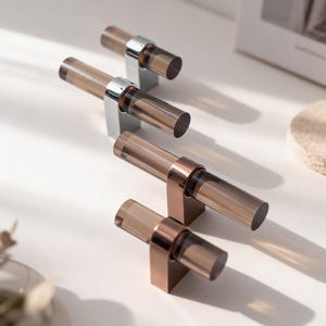 May include: Four T-shaped cabinet knobs featuring translucent brown cylindrical bars. Two knobs have polished chrome bases, while the other two have metallic copper-toned bases. Each knob consists of a horizontal bar mounted on a vertical support.