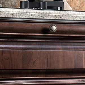 May include: Close-up of a dark brown wooden cabinet drawer with an antique-style knob. The drawer features a detailed wood grain and a curved design. The knob is a muted gold colour.