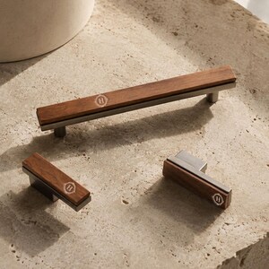 May include: A set of three cabinet pulls featuring a combination of dark brown wood and brushed metal. Each piece has a rectangular wooden inlay with a small circular emblem containing the letter H centred on the wood surface.
