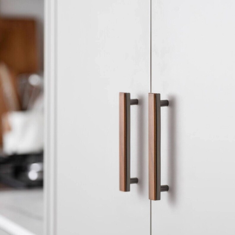 May include: Close-up of a white cabinet door featuring two bronze and wood-tone bar pulls. The pulls have a rectangular shape with a dark bronze base and a lighter wood-tone centre. The cabinet door has a clean, modern design.