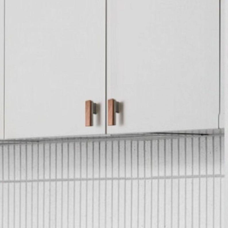 May include: Image of white kitchen cabinet doors with two pairs of copper-coloured rectangular cabinet pulls. The pulls are vertically aligned and have a simple, modern design. A white and grey tiled backsplash is in the background.