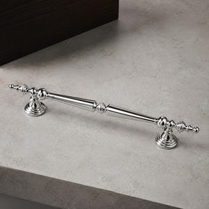 May include: A polished chrome cabinet pull featuring a long, slender, tapered central bar with decorative spherical and tiered finial accents at both ends, supported by two circular mounting bases with a reflective, metallic finish.
