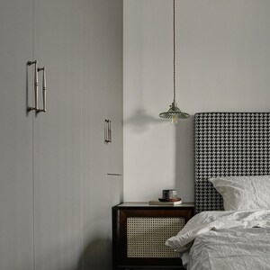 May include: A bedroom interior with a grey wardrobe and decorative handles, a bedside table, and a bed with a houndstooth headboard. A pendant light hangs above the nightstand. The bed has white linens.