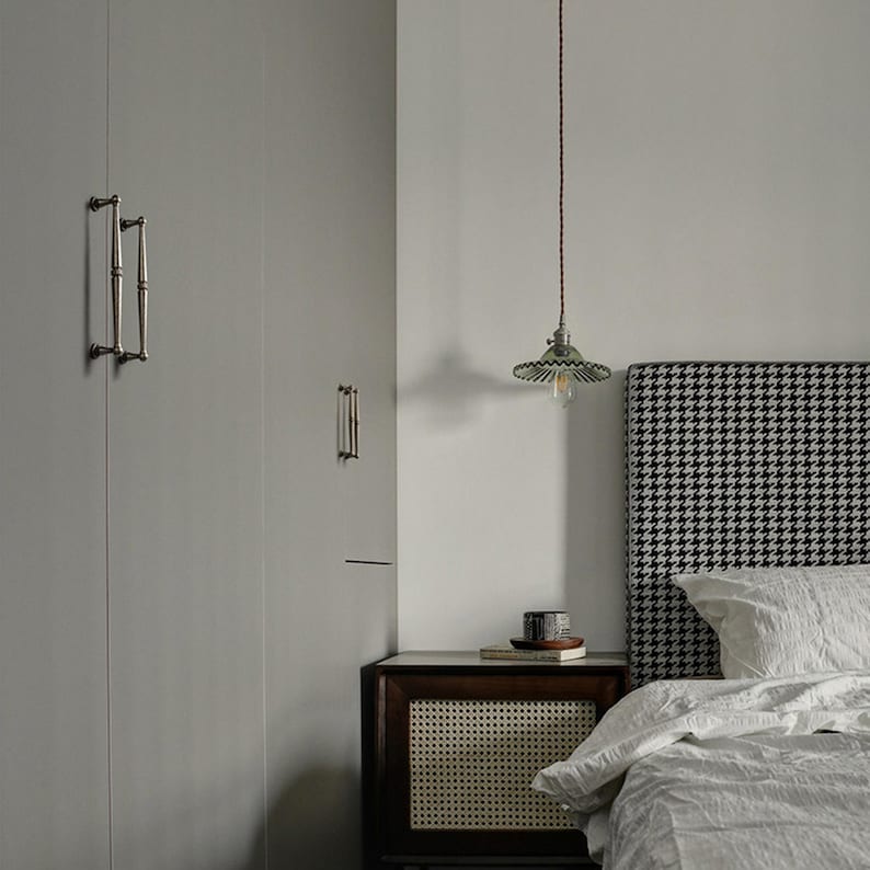 May include: A bedroom interior with a grey wardrobe and decorative handles, a bedside table, and a bed with a houndstooth headboard. A pendant light hangs above the nightstand. The bed has white linens.