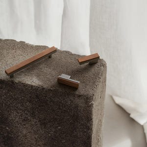 May include: Three pieces of modern cabinet hardware. The hardware features a combination of wood and metal. The largest piece is a long handle, while the other two are smaller knobs. The wood is a warm brown colour, and the metal is a cool silver.