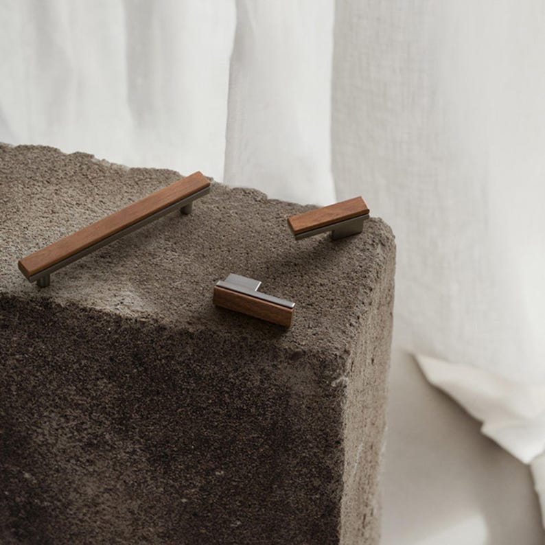 May include: Three pieces of modern cabinet hardware. The hardware features a combination of wood and metal. The largest piece is a long handle, while the other two are smaller knobs. The wood is a warm brown colour, and the metal is a cool silver.