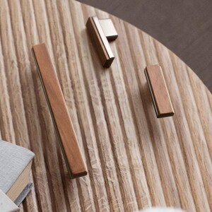 May include: A selection of contemporary cabinet handles. The handles have a rectangular wooden design with metallic detailing. The wood is a warm brown, and the metal is silver. They are arranged on a light brown, ridged surface.