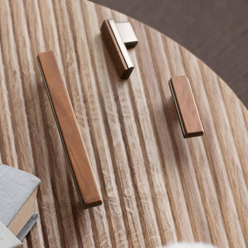 May include: A selection of contemporary cabinet handles. The handles have a rectangular wooden design with metallic detailing. The wood is a warm brown, and the metal is silver. They are arranged on a light brown, ridged surface.