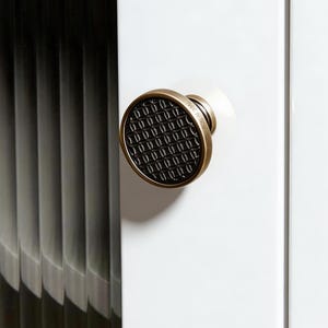 May include: A round cabinet knob with a brushed brass finish and a black center inlay featuring a repeating oval pattern. The edge of the brass rim is engraved with the text MADE BY TIKON. The knob is mounted on a white cabinet door surface.