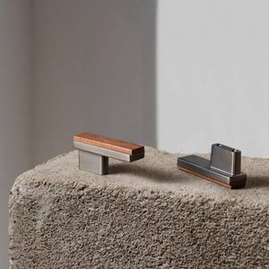 May include: Two contemporary cabinet pulls. One has a rectangular, brushed metal base with a brown wood top. The other has a similar design, with a brushed metal base and a copper-coloured accent. Both are displayed on a textured, grey surface.