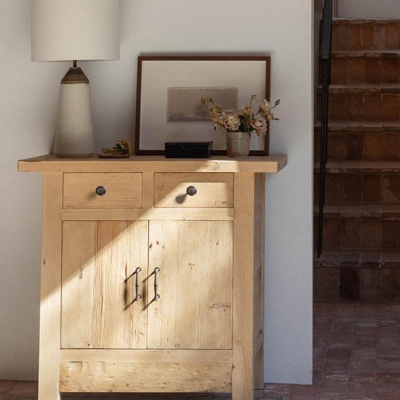 May include: A light-coloured wooden cabinet with two drawers and two doors. The cabinet has black hardware and is topped with a lamp, framed artwork, and a vase of flowers. A staircase is visible in the background.