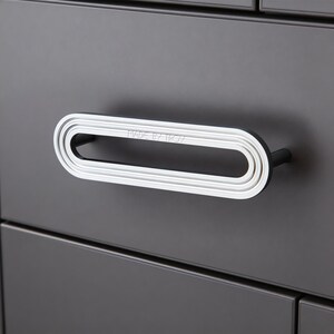 May include: A horizontal oval-shaped drawer pull with a polished silver finish and concentric grooved detailing. The center of the handle features the embossed text MADE BY TROY. The hardware is mounted on a matte dark gray cabinet drawer front.