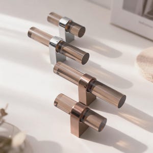 Peut inclure: Quatre poignées de meubles avec des poignées cylindriques en acrylique brun fumé. Deux poignées ont des bases en métal argenté et deux ont des bases en cuivre. Les poignées sont disposées sur une surface blanche, suggérant une esthétique moderne.