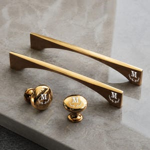 May include: Two gold-toned metal drawer pulls and two matching round cabinet knobs. Each piece features a polished finish and is engraved with the letter M centred above a small antler graphic. The hardware has a sleek, curved design.
