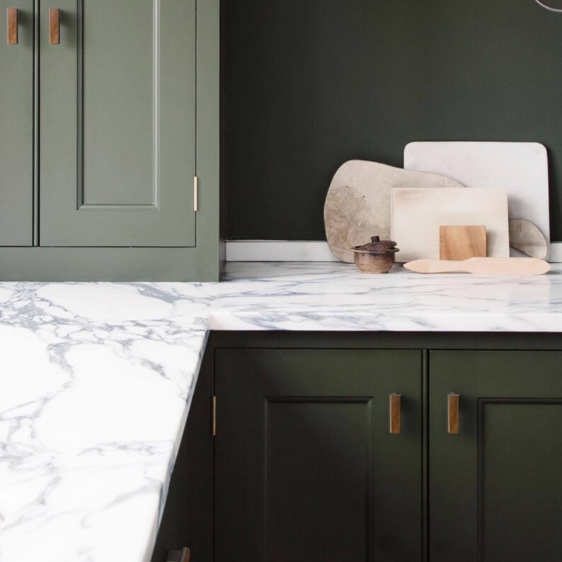 May include: A kitchen interior with olive green cabinets and wooden handles, a white marble worktop, and a dark green wall. Several wooden chopping boards and a small ceramic pot are displayed on the surface.