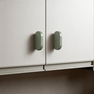 May include: Two sage green, oval-shaped cabinet knobs mounted on light gray cupboard doors. Each knob features a smooth, rounded profile with the text MADE IN ITALY embossed vertically on the side of the right-hand handle, providing a functional grip.