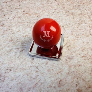 Op de afbeelding: Een glanzend rood bolvormig object met een witte letter M en een witte gewei-afbeelding, gemonteerd op een vierkante, reflecterende zilverkleurige metalen voet. Het oppervlak van de bol is glad en reflecteert het licht, wat het monogram benadrukt.