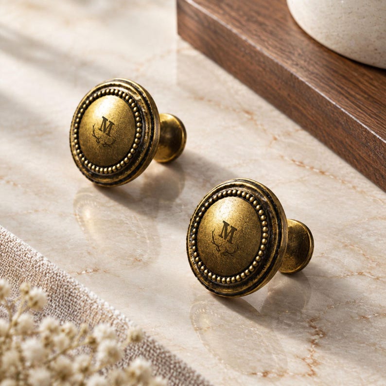 May include: Two round cabinet knobs with an antique brass finish. Each knob features a raised beaded border and a central engraving of the letter M positioned above a small antler graphic. The hardware has a weathered, metallic texture.