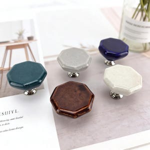 以下が含まれることがあります： 様々な色のセラミック製キャビネットノブのコレクション。ティール、グレー、ブラウン、ネイビーブルー、オフホワイトなど。各ノブは八角形で、銀色のベースが付いています。家具や家の装飾プロジェクトに適しています。